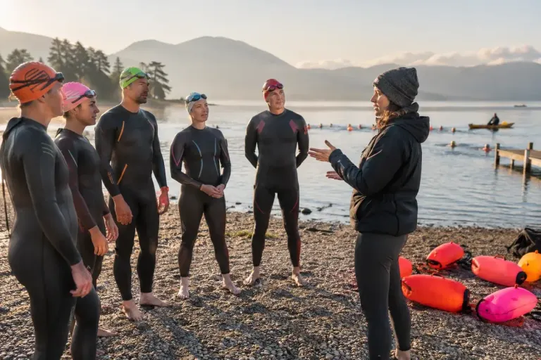 Adult swimmers listening to a coach beside a supervised open-water swimming area, bright tow floats on shore.