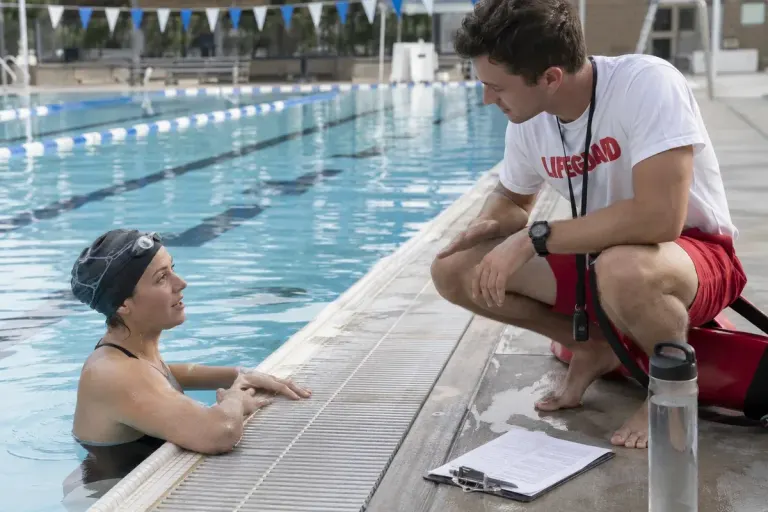 Adult swimmer speaking to a lifeguard beside a public pool with clearly visible lane markings and pool floor, calm daylight.