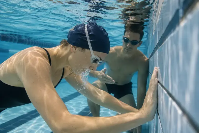 An adult swimmer practising gentle underwater exhalation at a pool wall with a coach nearby.